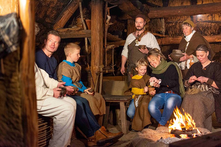 Besucher in historischem Gewandt vor einer Feuerstelle in einer Hütte